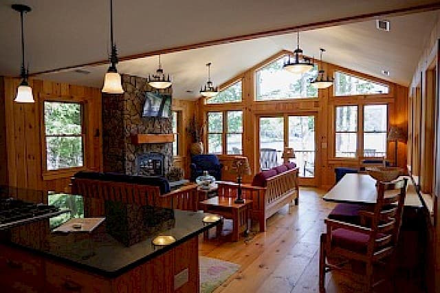 A warm cabin great room with a vaulted ceiling, wood paneling, a stone fireplace, and a wall of glass doors opening to the outdoors.