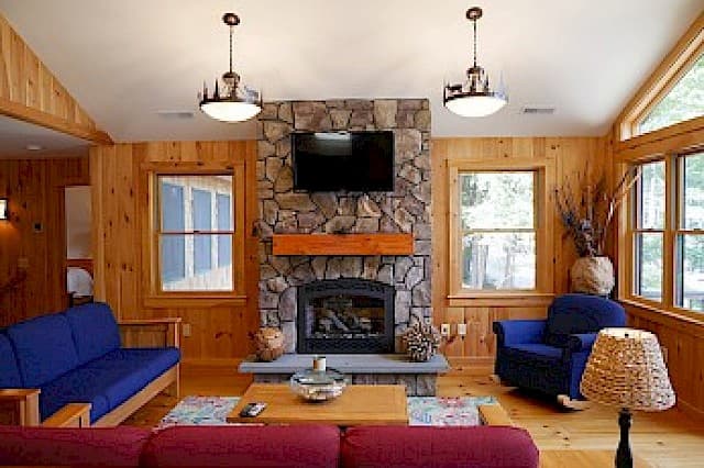 A warm cabin living room with wood paneling, a vaulted ceiling, a stone fireplace with a mounted TV, and blue and red seating.