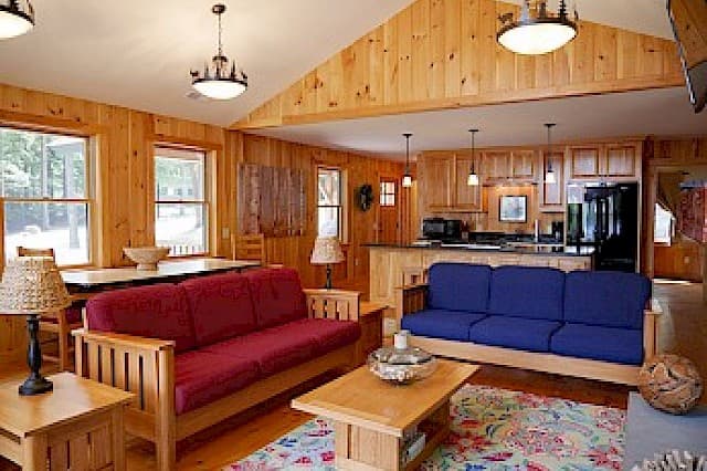 A bright cabin great room with a vaulted ceiling, wood paneling, two futon sofas, a dining area, and a functional kitchen.