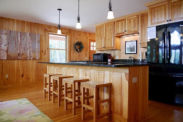 A bright cabin kitchen with light wood paneling, hardwood floors, a breakfast bar with four stools, and black countertops.