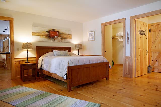 A bright cabin bedroom with hardwood floors, a dark wood bed, two lamps, and an open doorway leading to a bathroom and closet area.