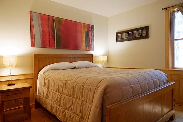 A bright cabin bedroom with a light wood bed frame, two lamps, and a large abstract painting with red and yellow tones above the bed.