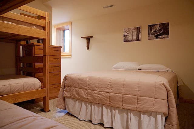 A simple cabin bedroom featuring a double bed and a wooden bunk bed, with light wood furniture, two prints, and a small window.