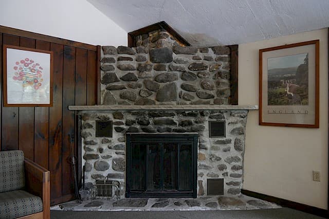 A large, rustic stone fireplace with a dark metal insert, flanked by wood paneling and two framed art prints in a cabin interior.