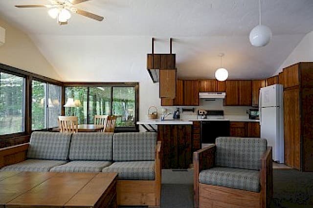 An open-plan cabin great room with a vaulted ceiling, featuring a living area with a plaid sofa and an open-concept kitchen with wood cabinets.