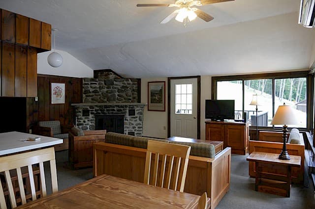 A large cabin great room with a vaulted ceiling, wood paneling, a stone fireplace, and a dining area in the foreground.