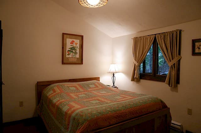 A cozy cabin bedroom with a vaulted ceiling, a bed with a geometric quilt, a bedside lamp, and a curtained window.