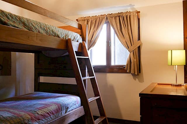 A simple cabin bedroom featuring a wooden bunk bed with a ladder, a small window with beige curtains, and a bedside lamp.