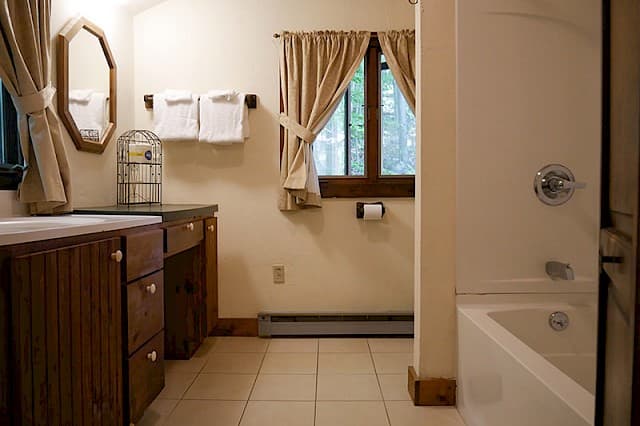 A cabin bathroom with a double vanity area, a white bathtub on the right, and a window with beige curtains over the toilet area.