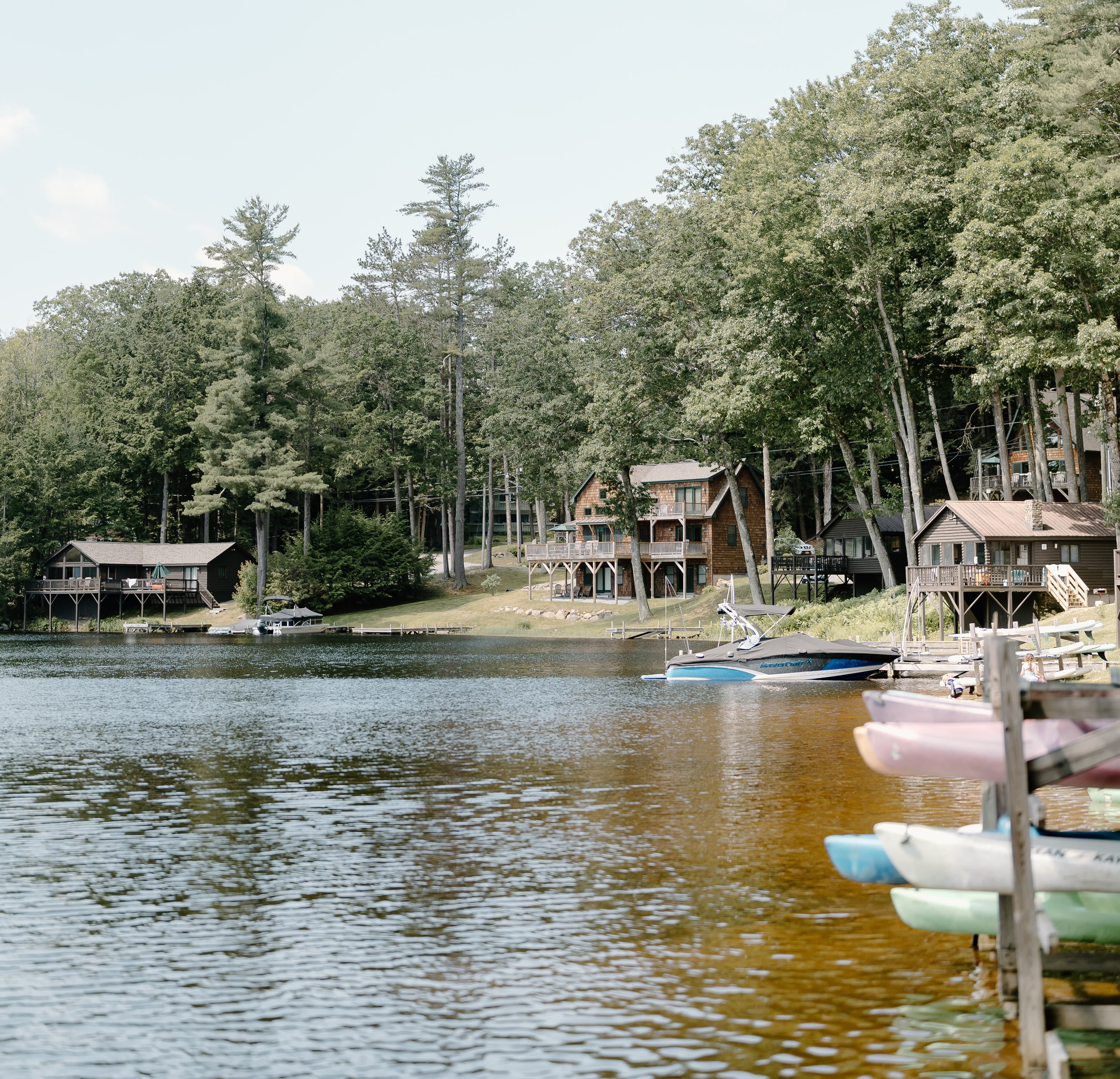 A serene lake scene featuring wooden cabins and colorful kayaks along the shore, surrounded by tall trees.