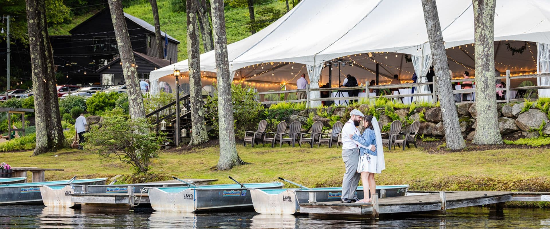 A couple embraces by the water in front of a large tent, surrounded by greenery and boats.