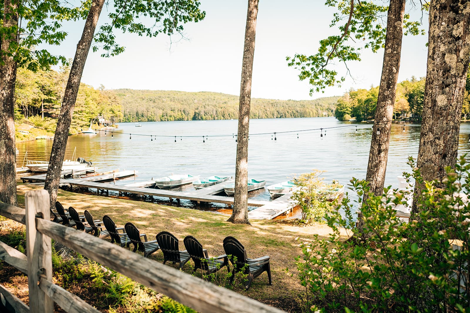 A serene lakeside scene featuring docked boats, lush trees, and chairs lined up along the shore.