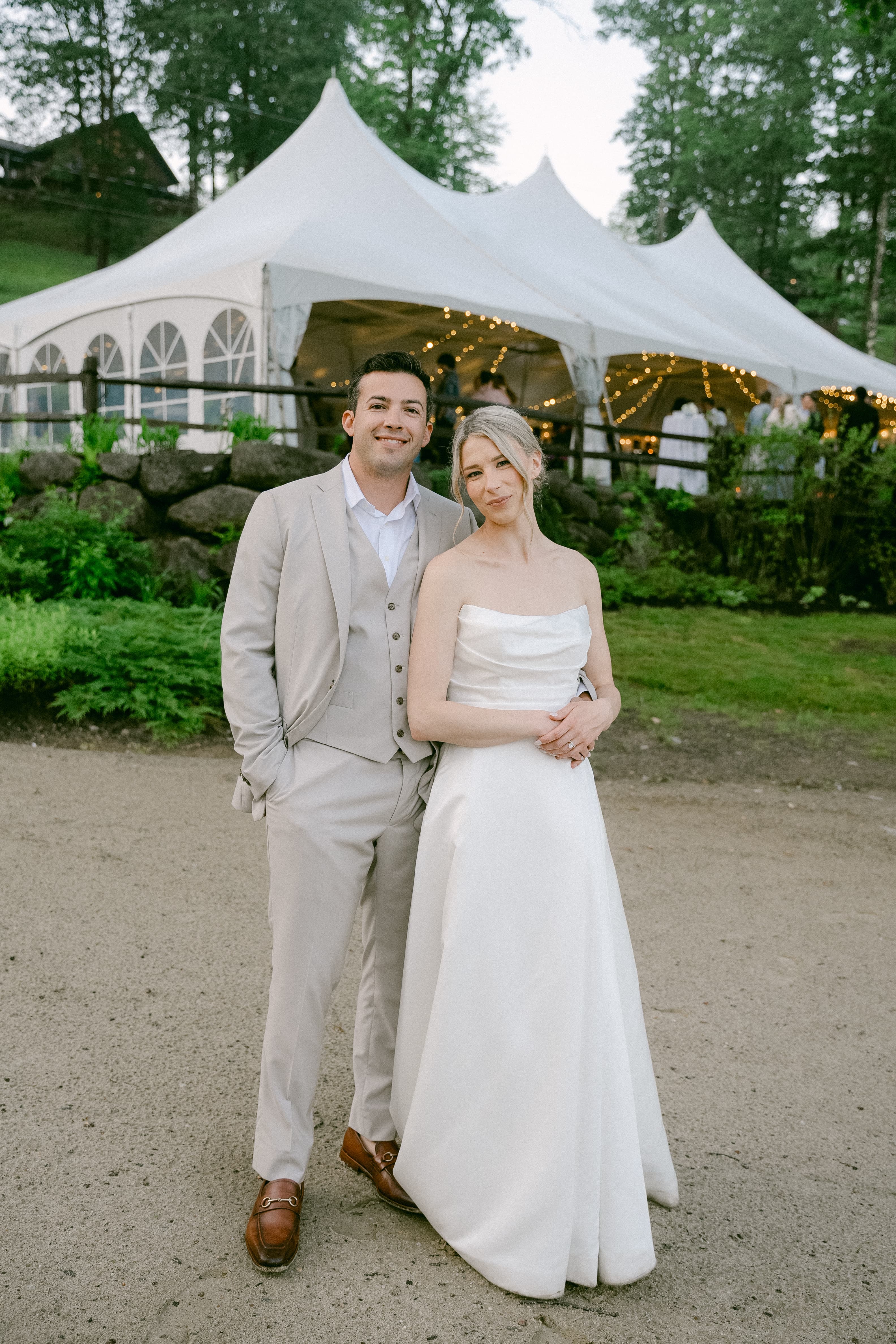 A bride and groom pose together in front of a tented outdoor venue.