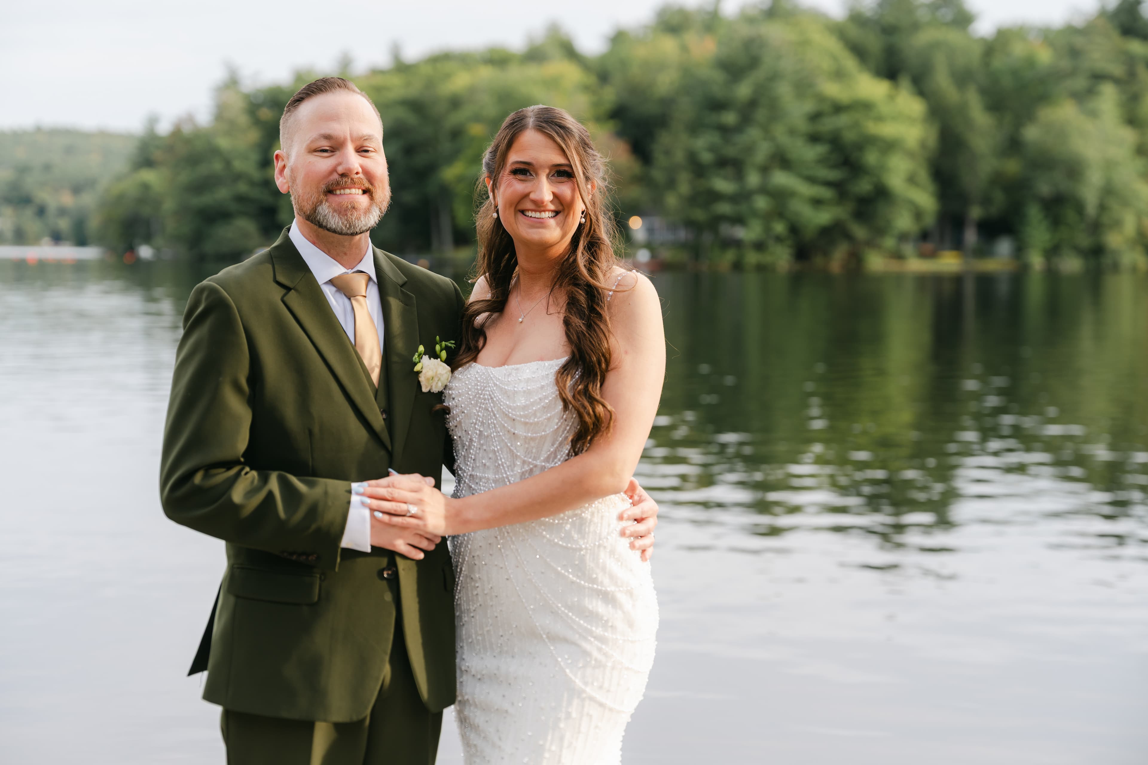 A smiling couple stands together by a serene lake, surrounded by greenery.