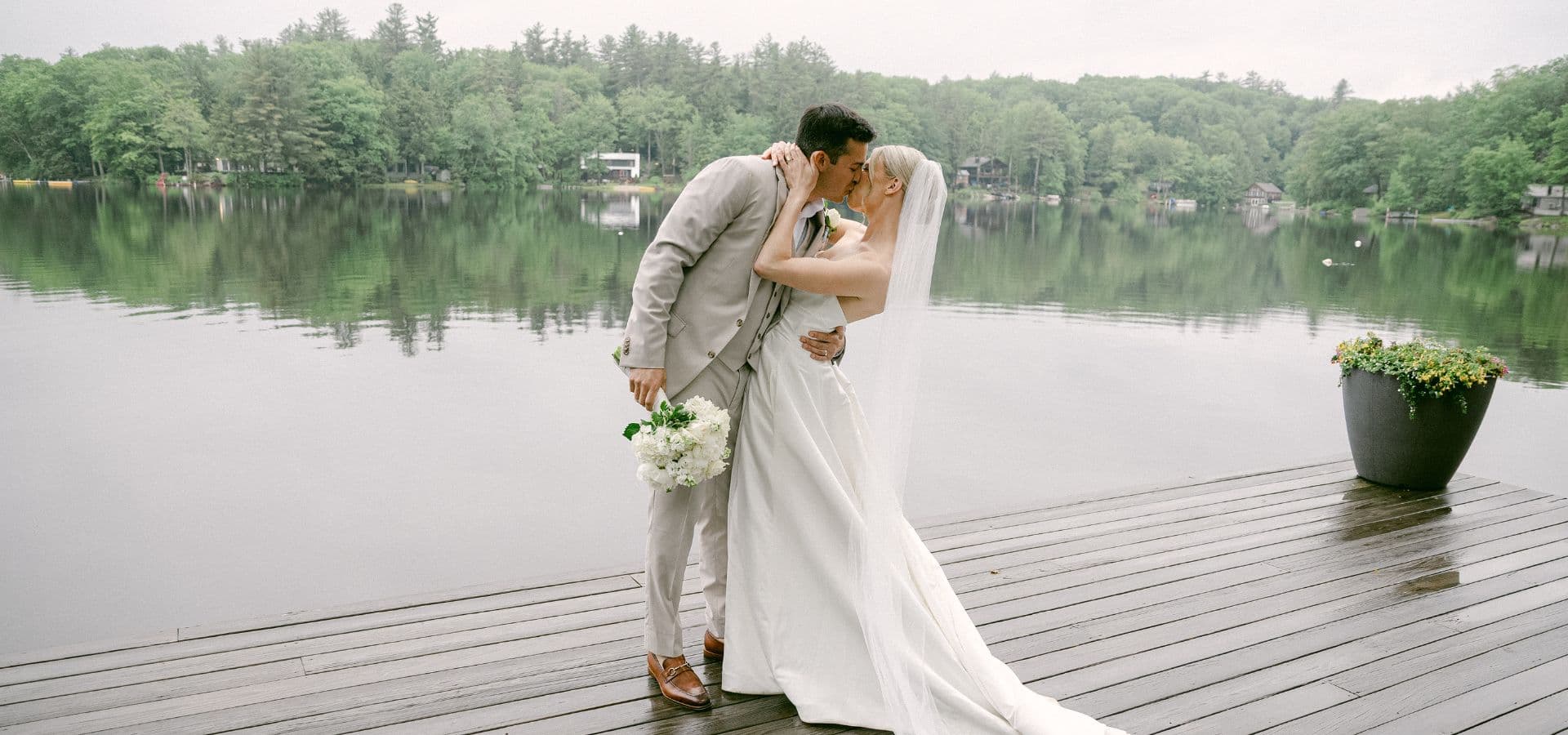 A bride and groom share a kiss on a dock by a serene lake, surrounded by lush greenery.