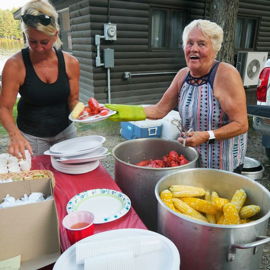 Two women prepare and serve food at a outdoor gathering, with lobsters and corn in large pots.