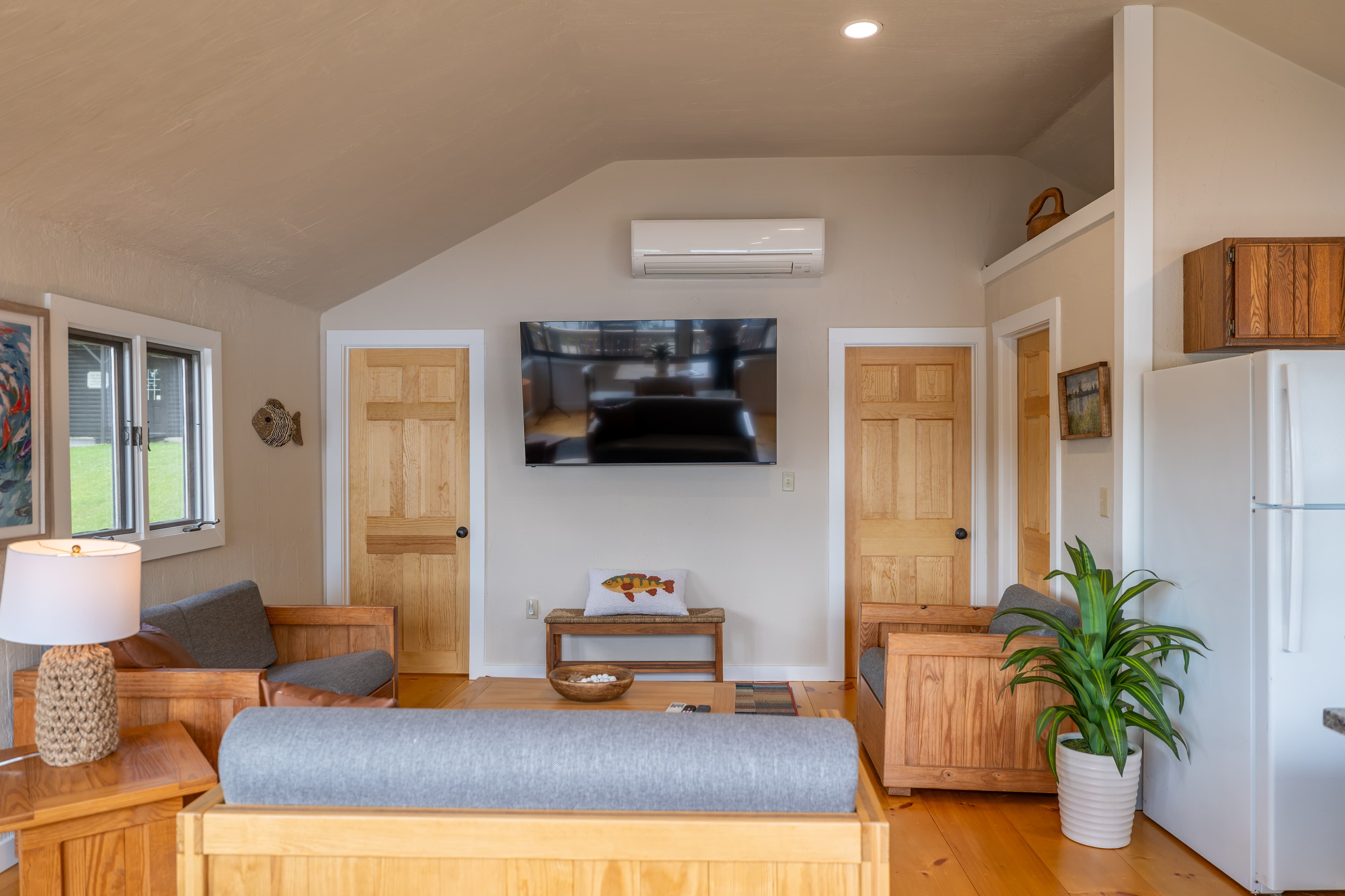A cozy cabin living room with a vaulted ceiling, natural wood furniture and a sofa, featuring a large stone fireplace and a mini-split AC unit.