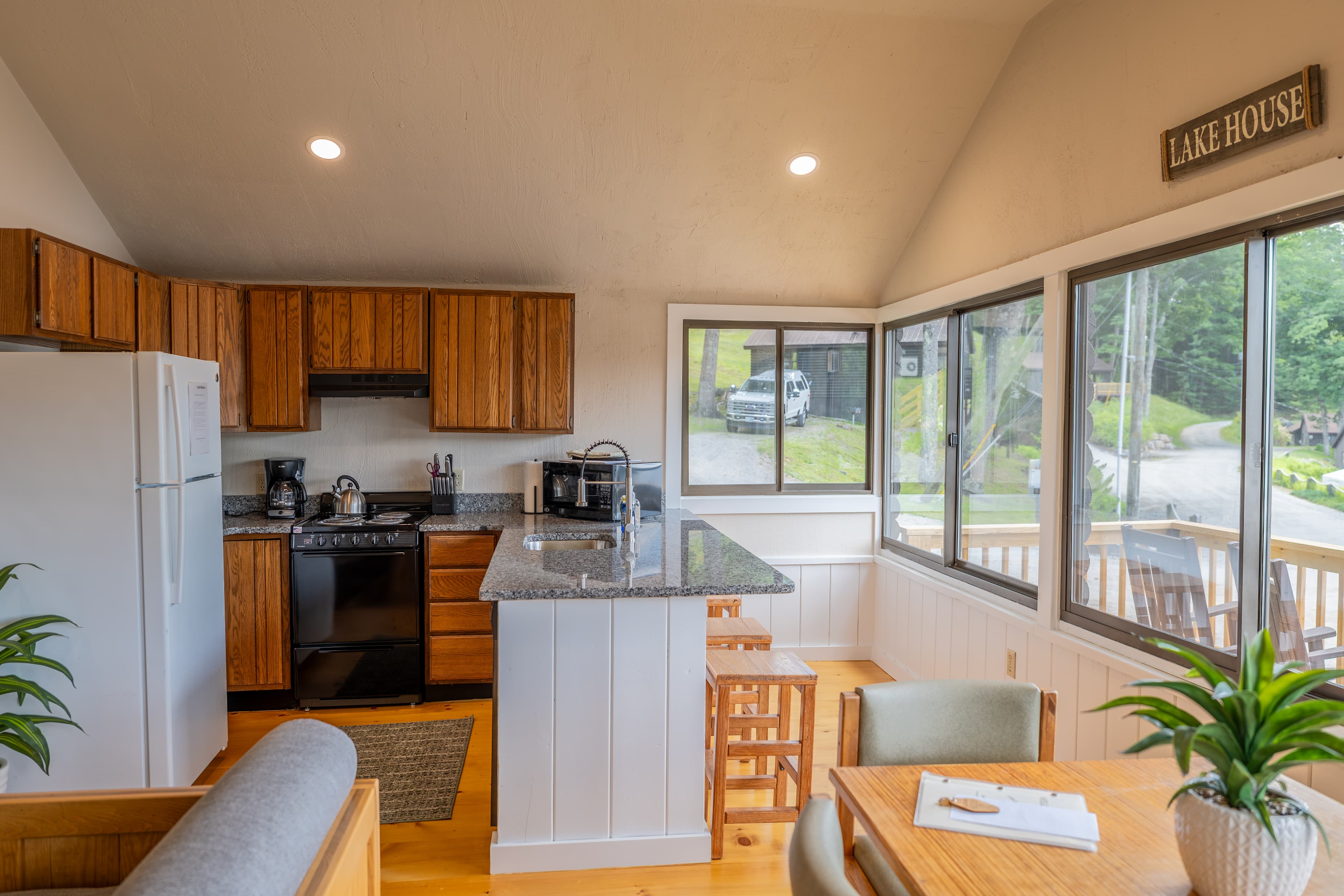 A cabin kitchen and dining area with a vaulted ceiling, wood cabinets, white appliances, and a dining nook next to large windows.