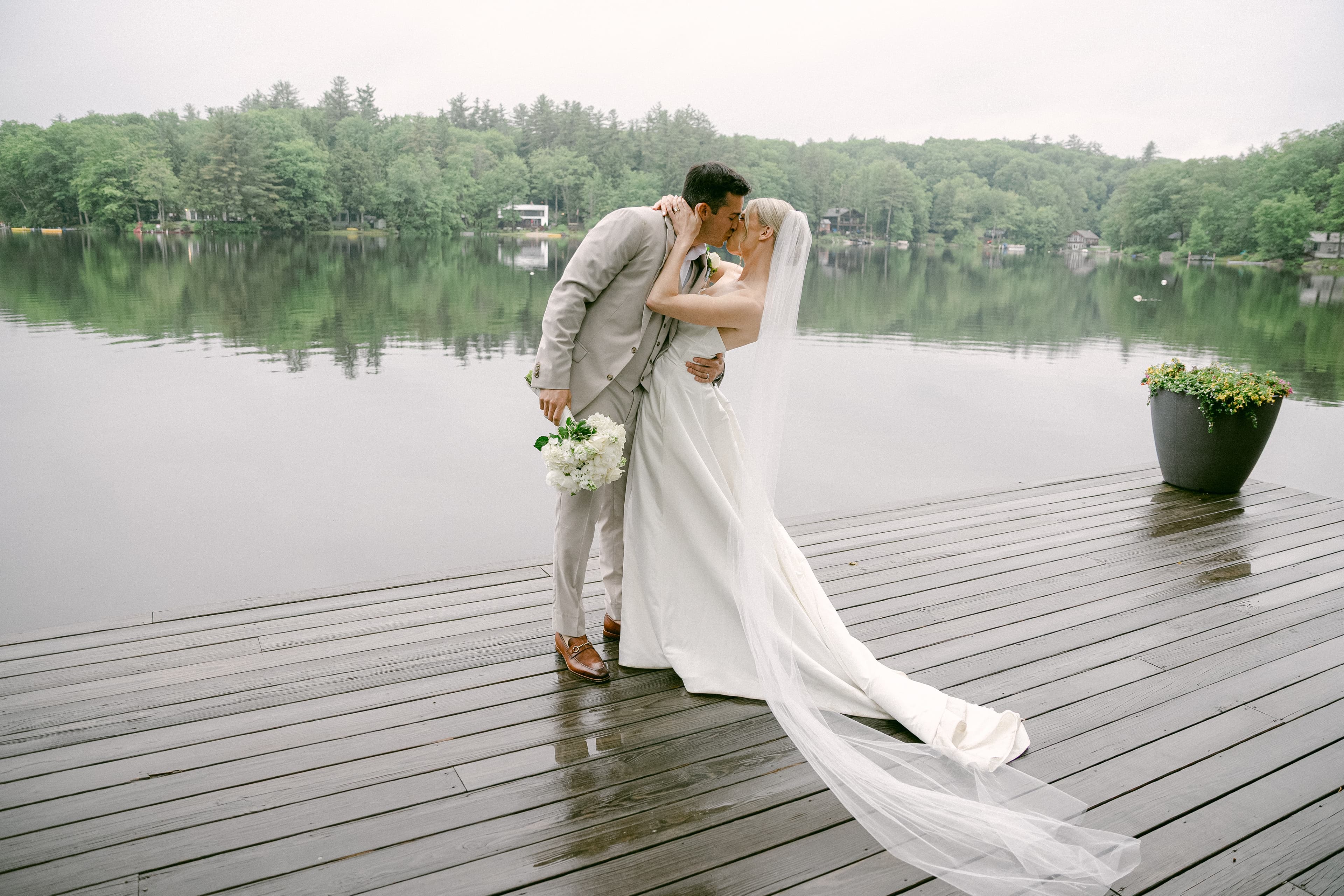 A bride and groom kiss on a wooden dock by a serene lake surrounded by greenery.