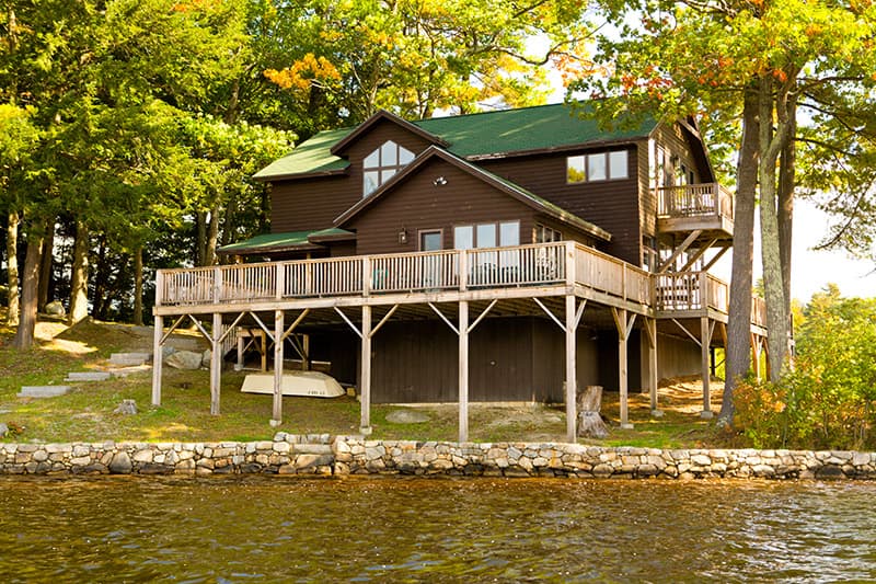 A two-story wooden cabin with a green roof, elevated on stilts beside a lake, surrounded by trees.