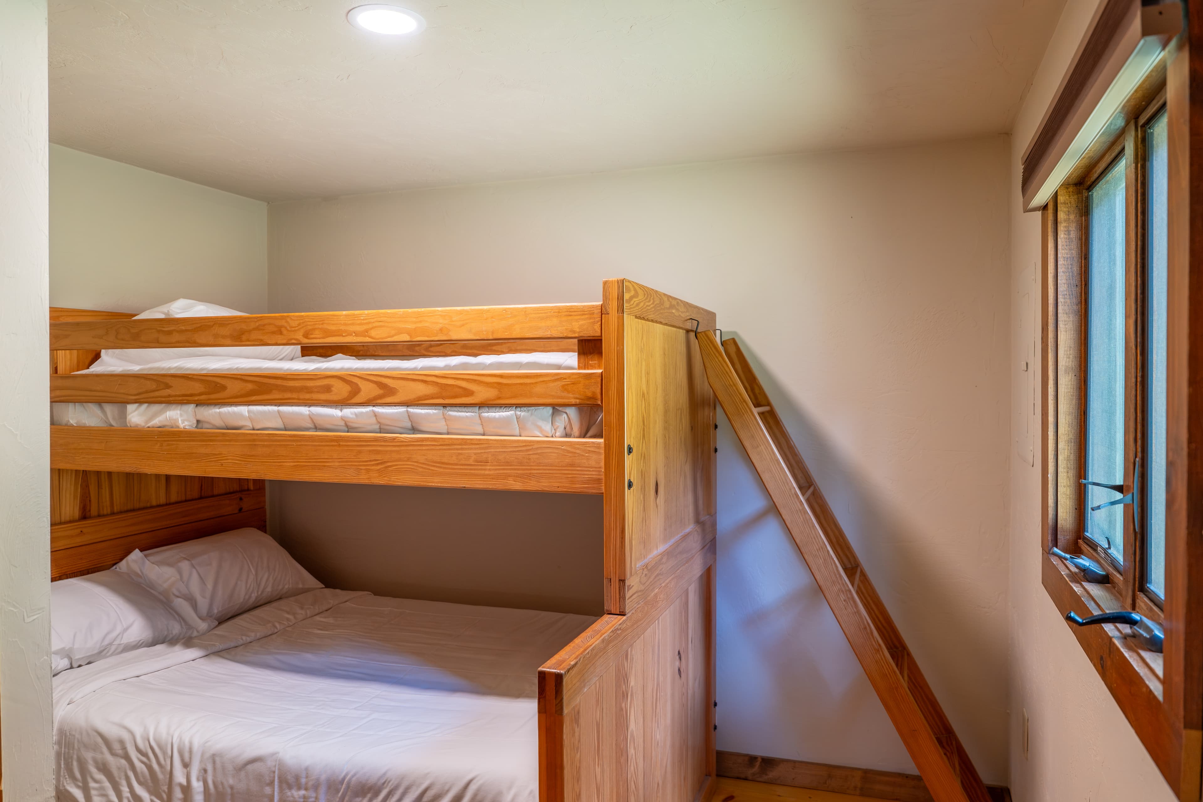 A simple cabin bedroom featuring natural wood bunk beds with a ladder, a small window with beige curtains, and two black and white prints.