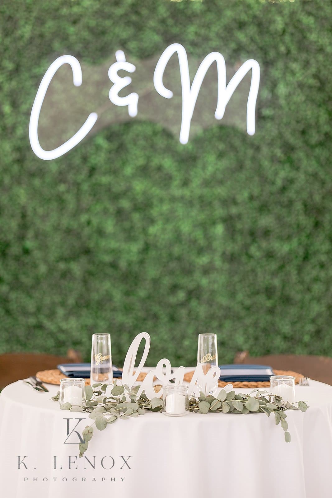 A decorated table featuring a "love" sign, glasses, and eucalyptus on a backdrop of greenery and illuminated initials.