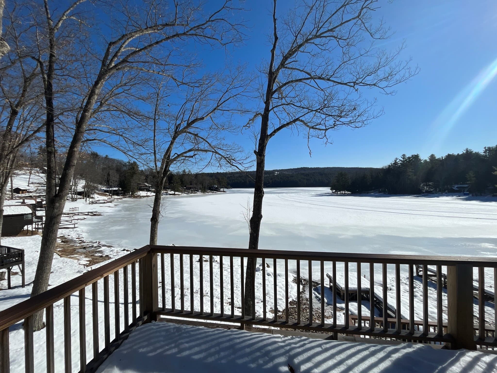 A snowy landscape with a frozen lake and clear blue sky viewed from a wooden deck.