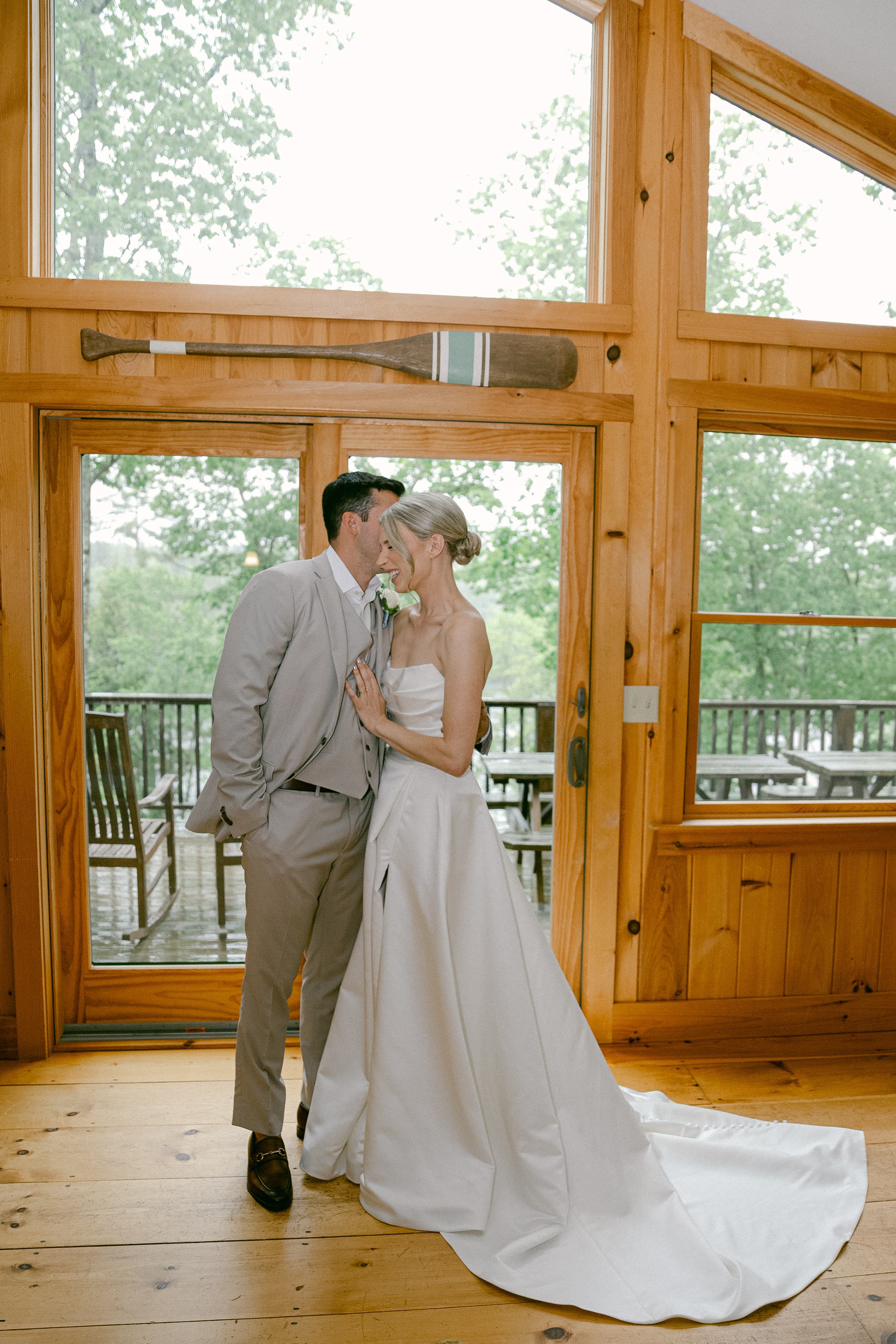 A bride and groom share a tender kiss indoors beside a large window overlooking greenery.