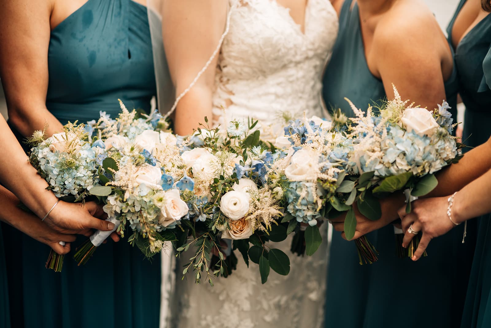 Bridesmaids in teal dresses hold elegant bouquets of blue and white flowers.