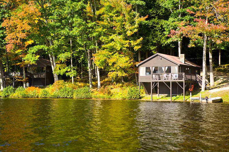Two cabins sit along a serene lake surrounded by vibrant autumn foliage.