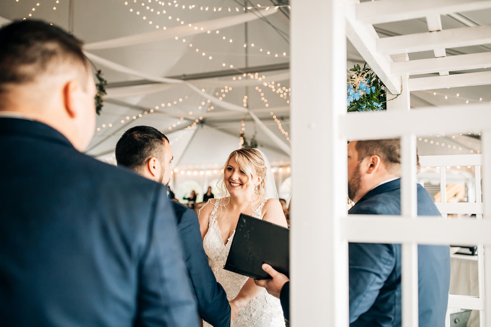 A smiling bride exchanges vows with a groom during a wedding ceremony under string lights.