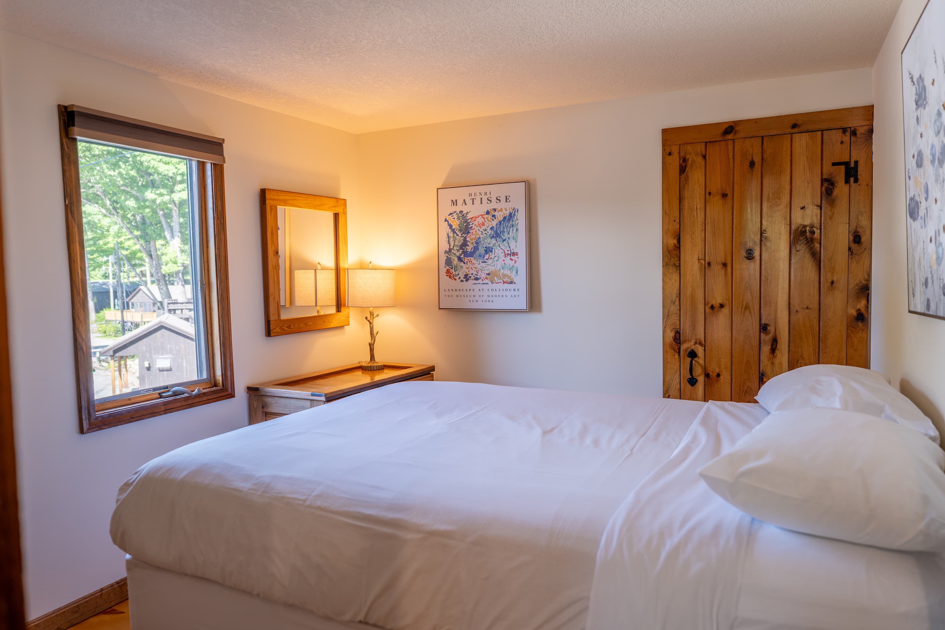 A cozy bedroom featuring a double bed with a colorful patterned quilt, a bedside table with a lamp, and a single window with beige curtains.