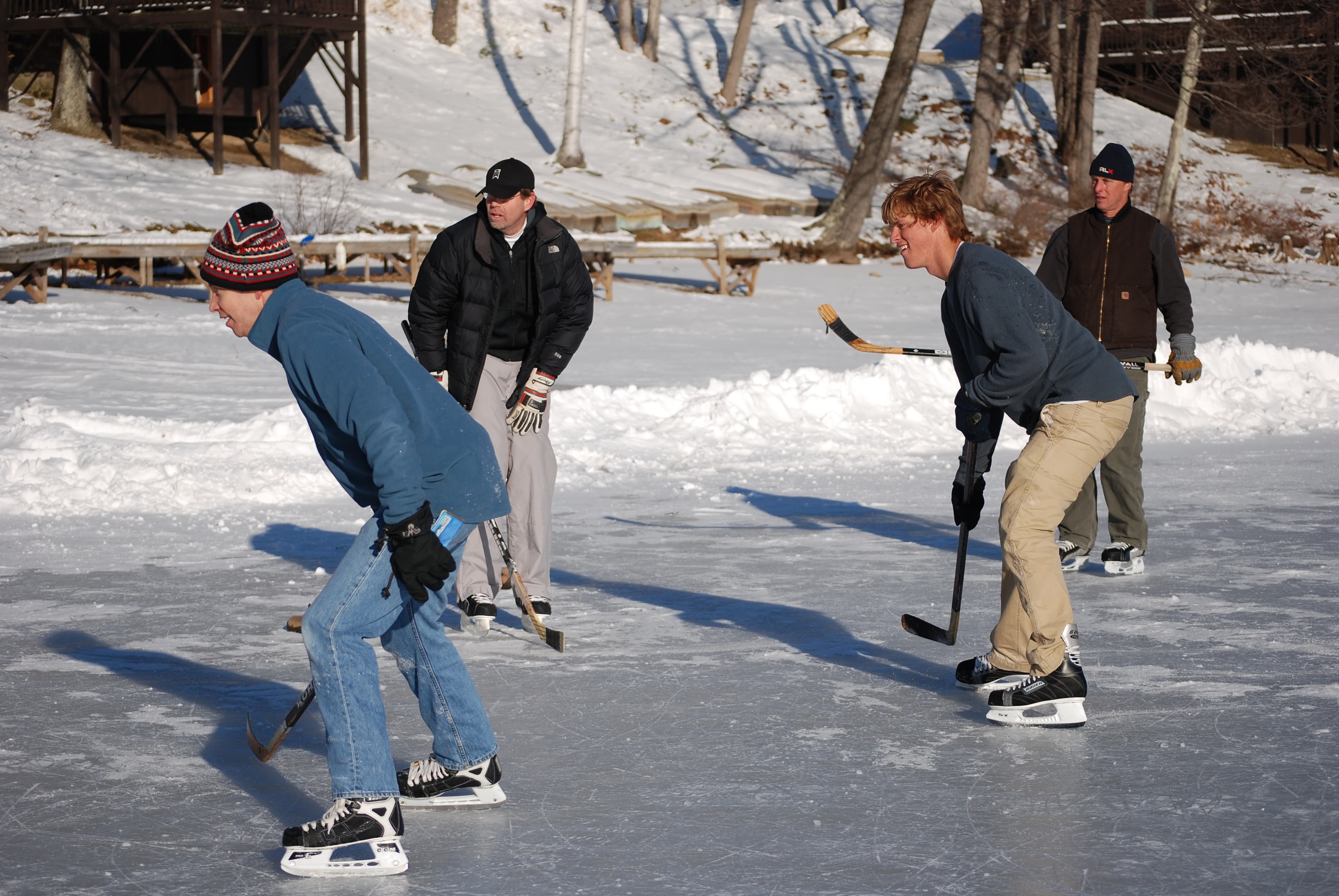 Four individuals are ice skating and playing hockey on a frozen lake surrounded by snow-covered trees.