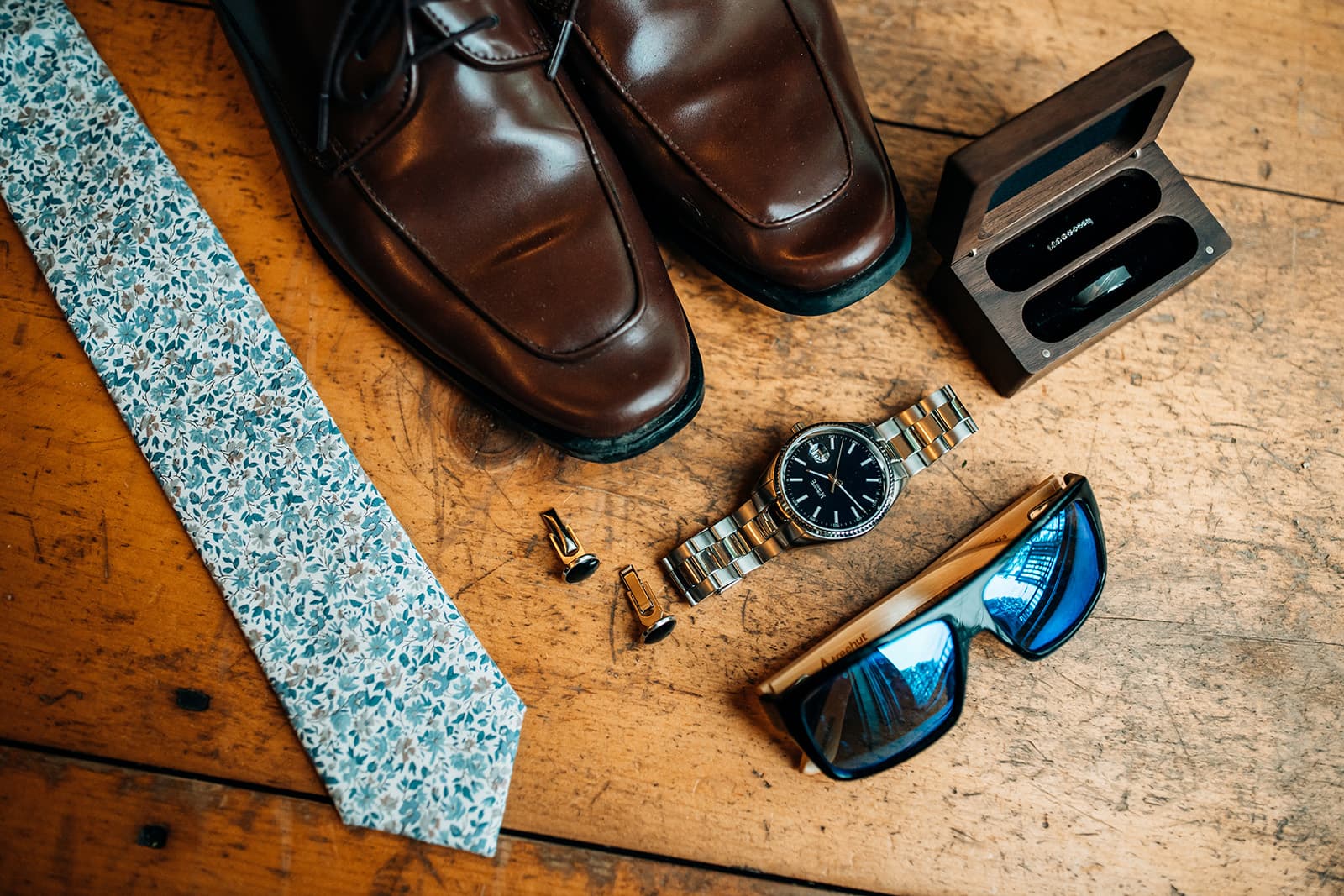 A flat lay arrangement of brown shoes, a floral tie, a watch, cufflinks, sunglasses, and a small wooden box on a wooden surface.