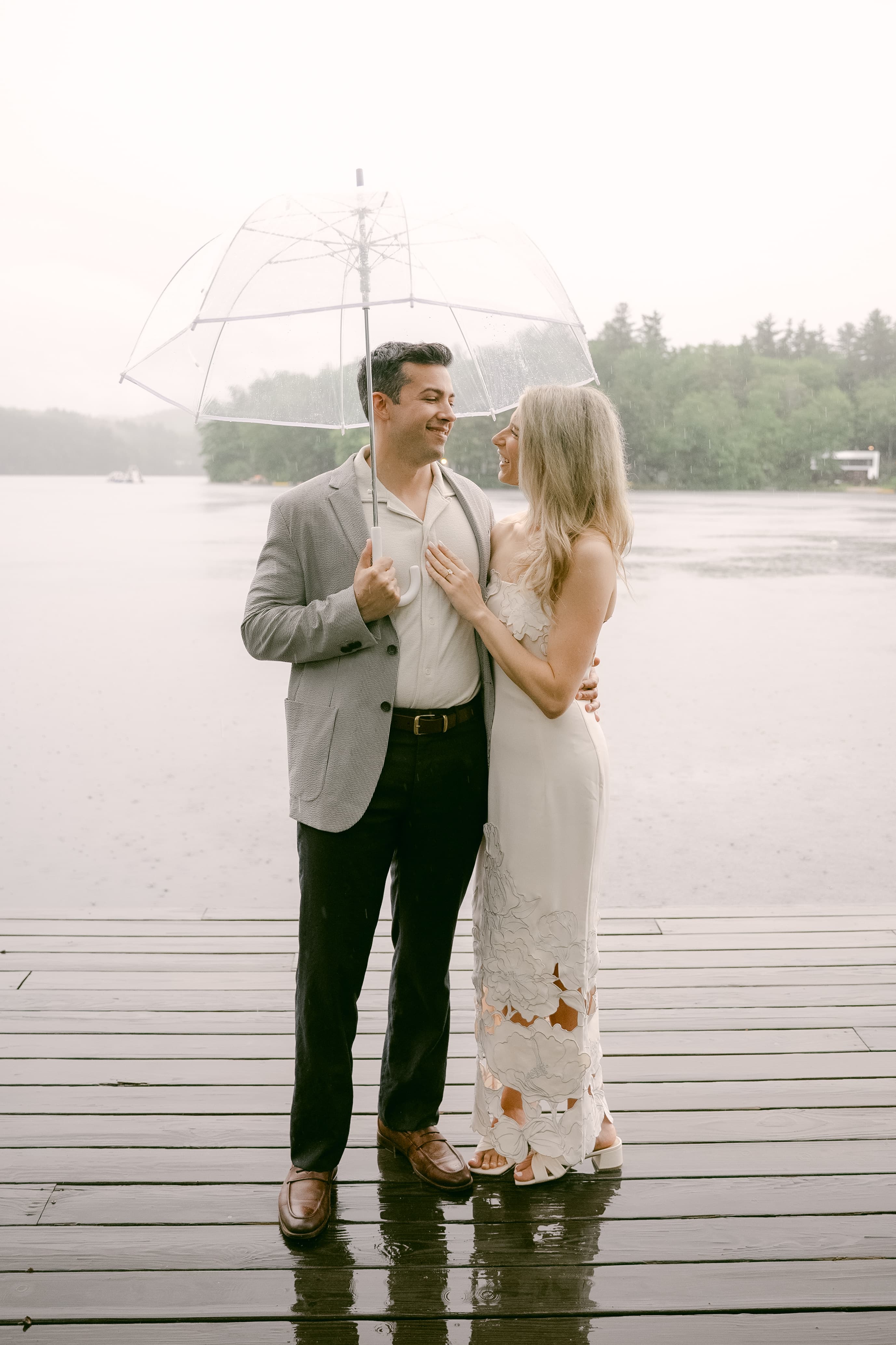 A couple stands under a clear umbrella on a rainy day by the water.