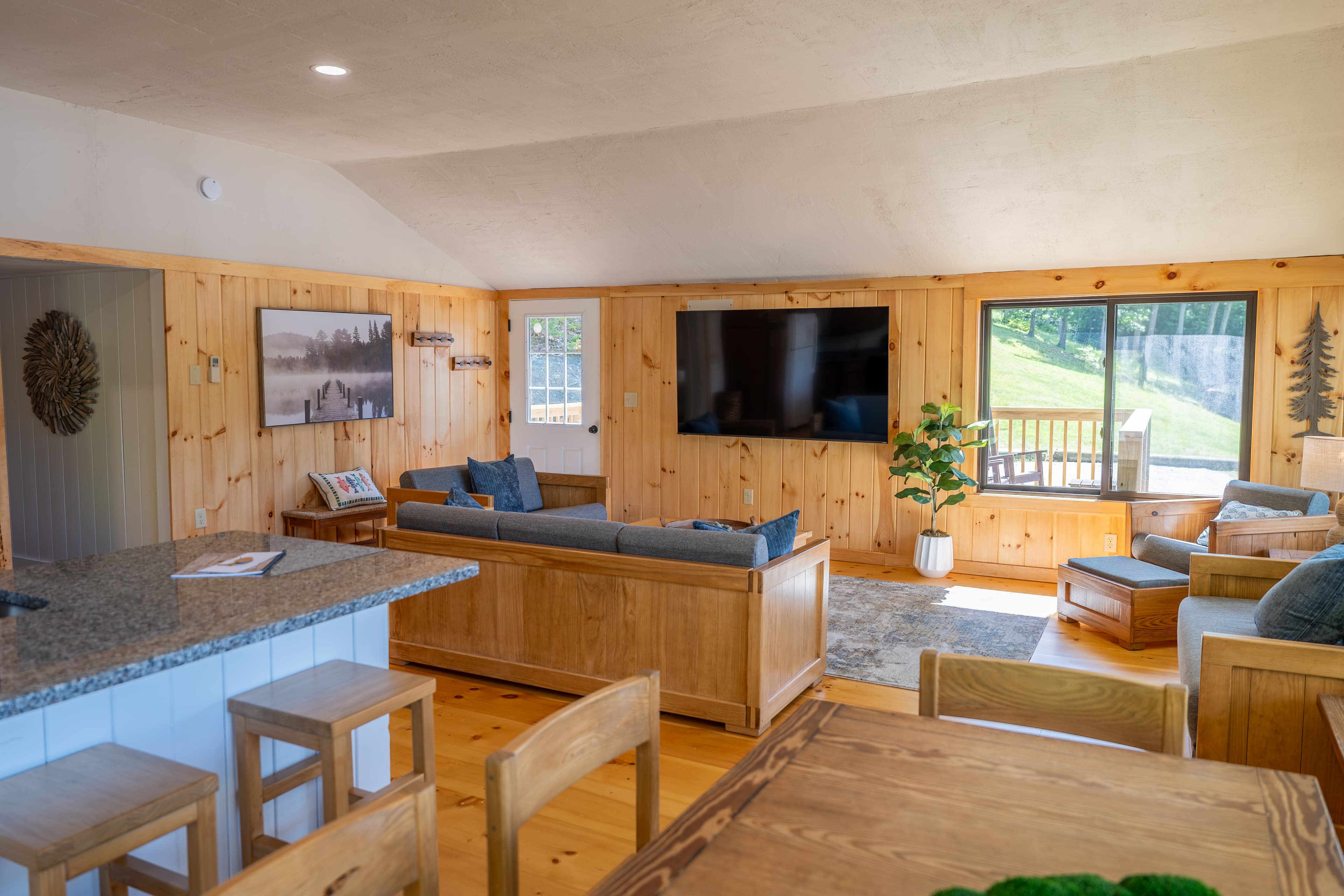 A large cabin great room with a vaulted ceiling, wood paneling, a stone fireplace, and a dining area in the foreground.