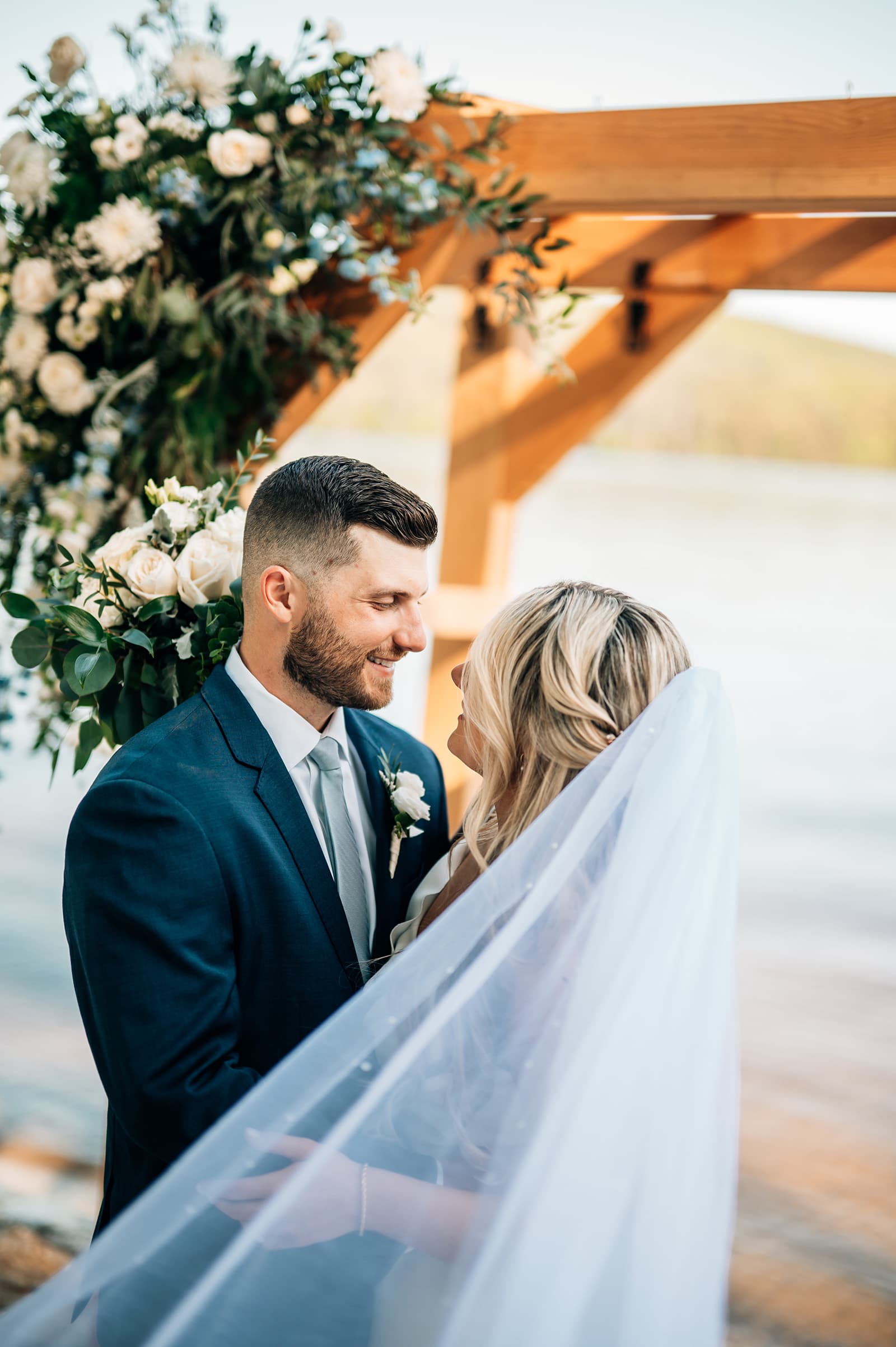 A couple gazes at each other under a floral arch by the water, sharing a romantic moment.