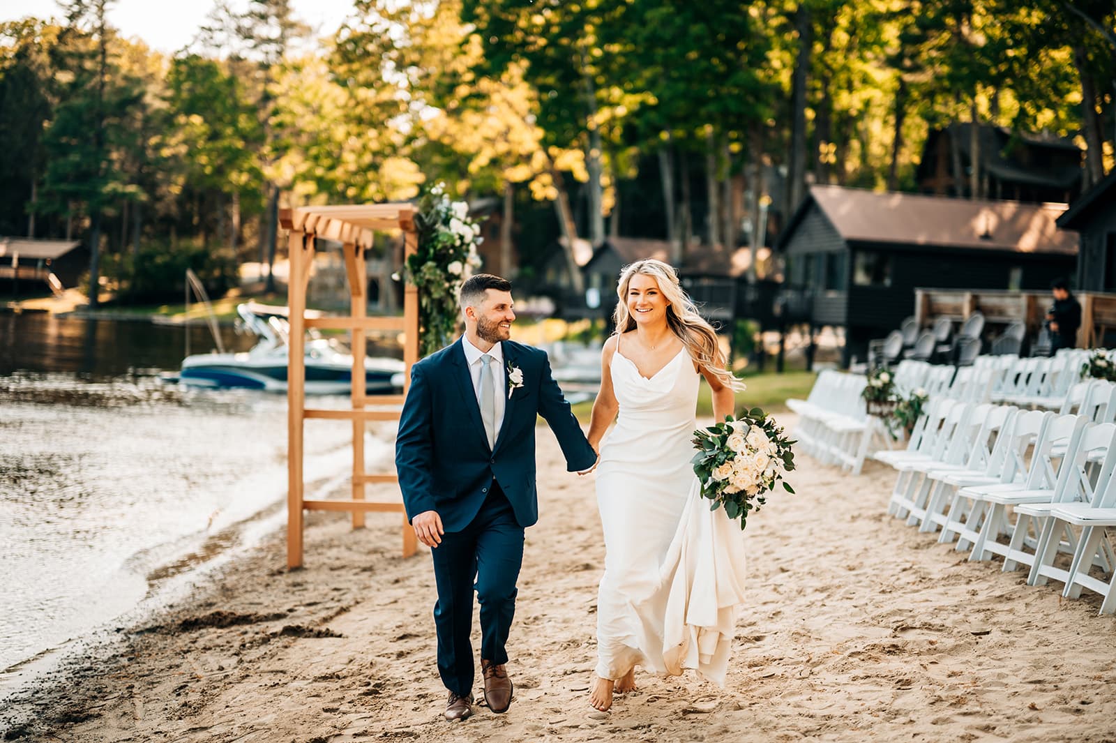 A bride and groom walk hand in hand along a sandy lakeside path, surrounded by greenery and wedding decor.