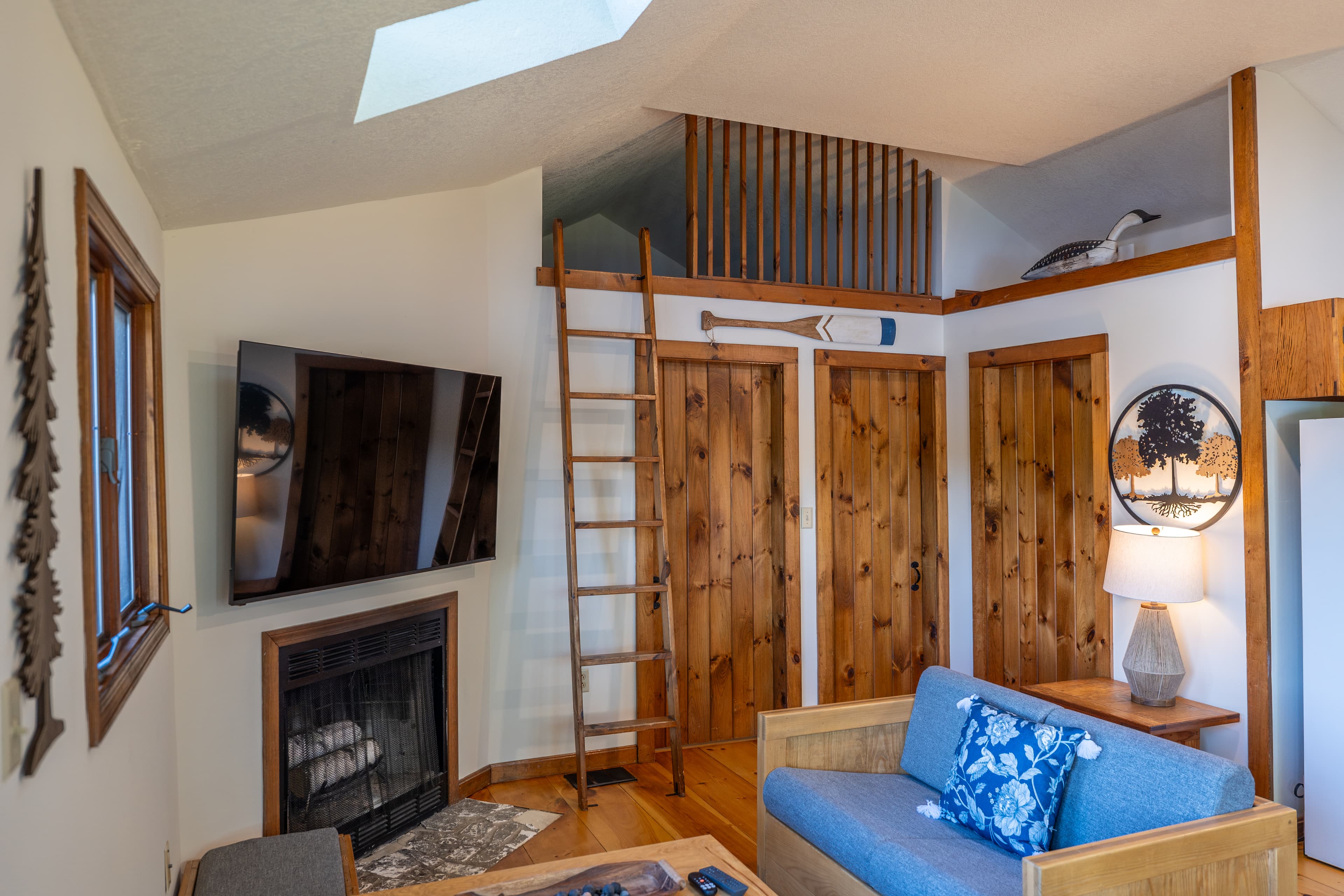 A bright cabin living area with a vaulted ceiling, hardwood floors, a blue futon, a small fireplace, and a ladder leading to a loft.