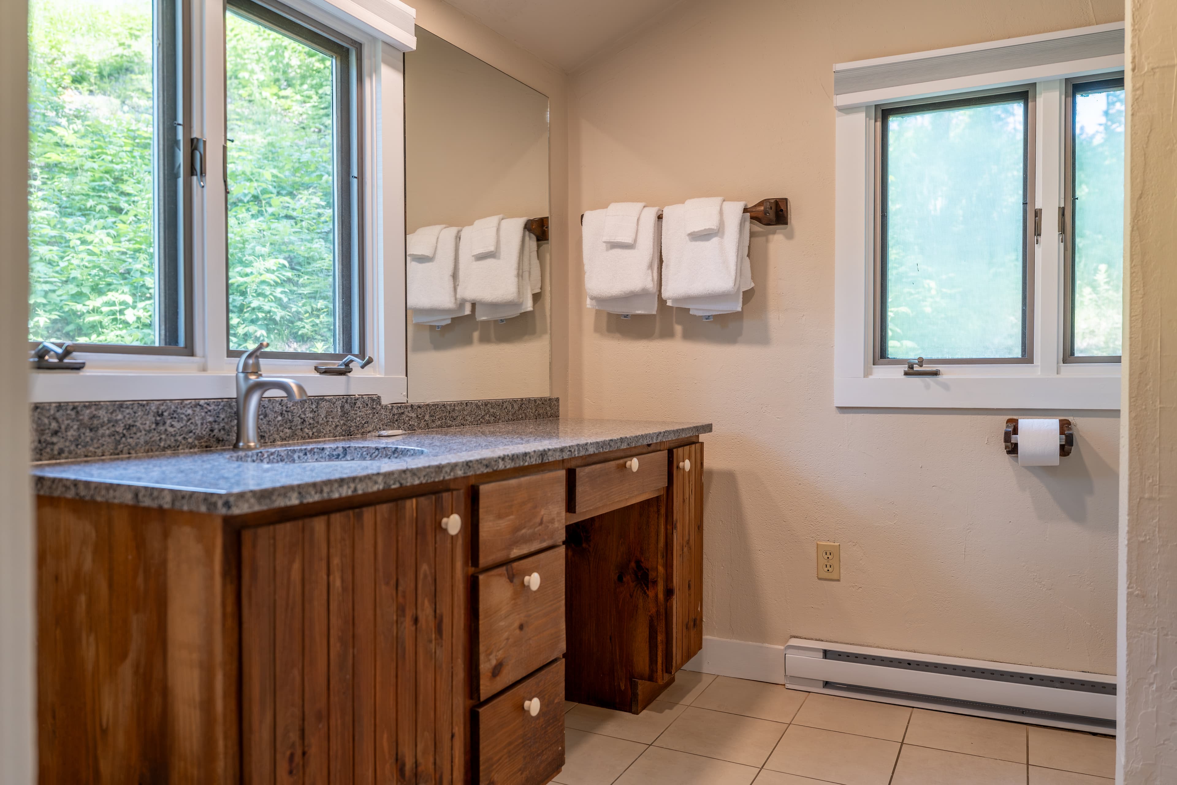 A cabin bathroom with a double vanity area, a white bathtub on the right, and a window with beige curtains over the toilet area.