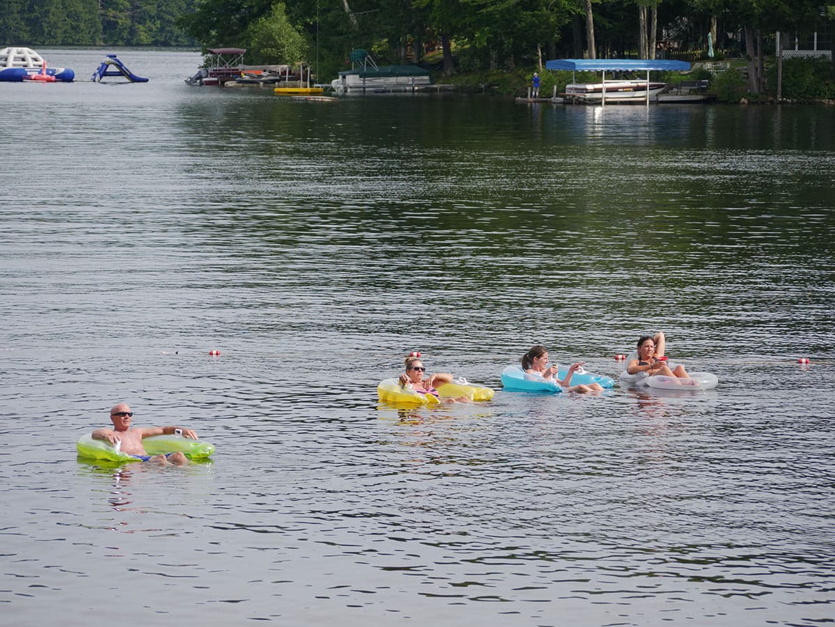 Five people relax on colorful inflatables in a calm lake surrounded by trees.