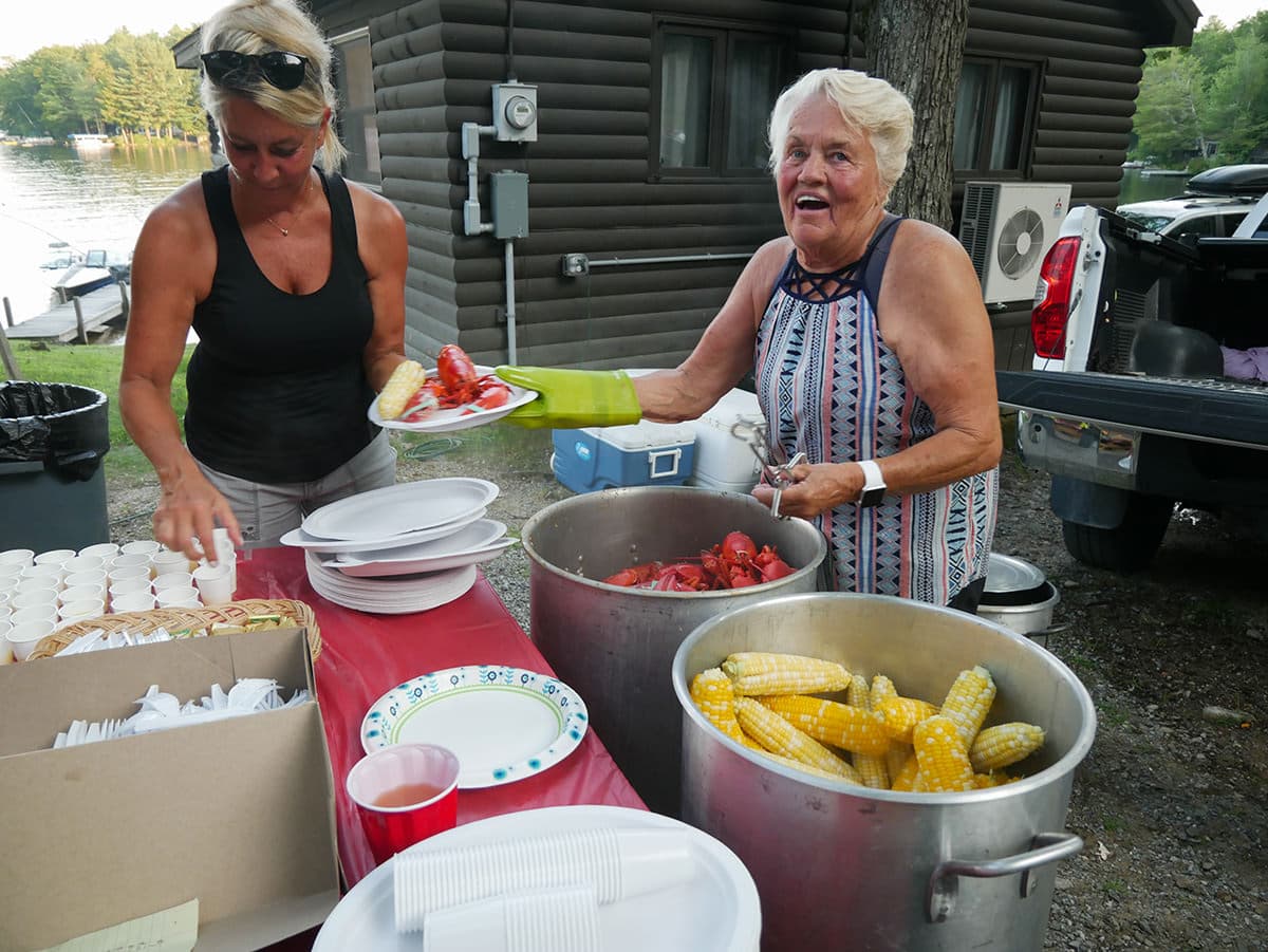 Two women prepare food outdoors, with one serving lobster and corn from pots while the other holds a plate.