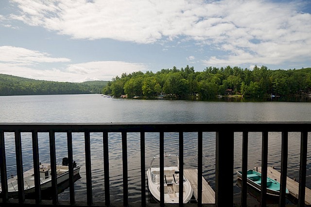 A wide view of a calm lake with a forested shoreline, seen from behind a dark railing, with a few small boats docked below.