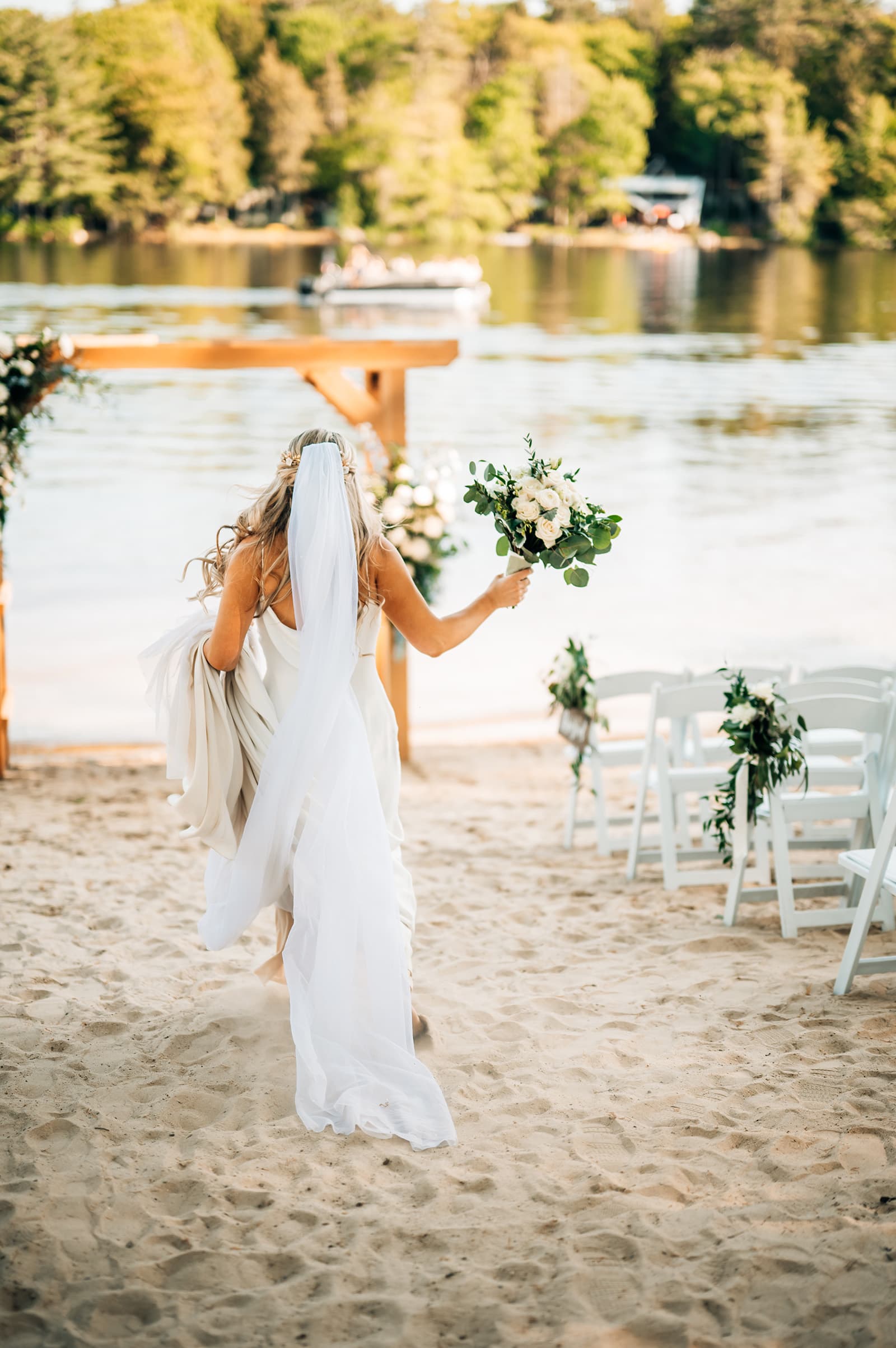 A bride walks towards a wooden arch by the water, holding a bouquet and her dress, with chairs arranged for guests nearby.