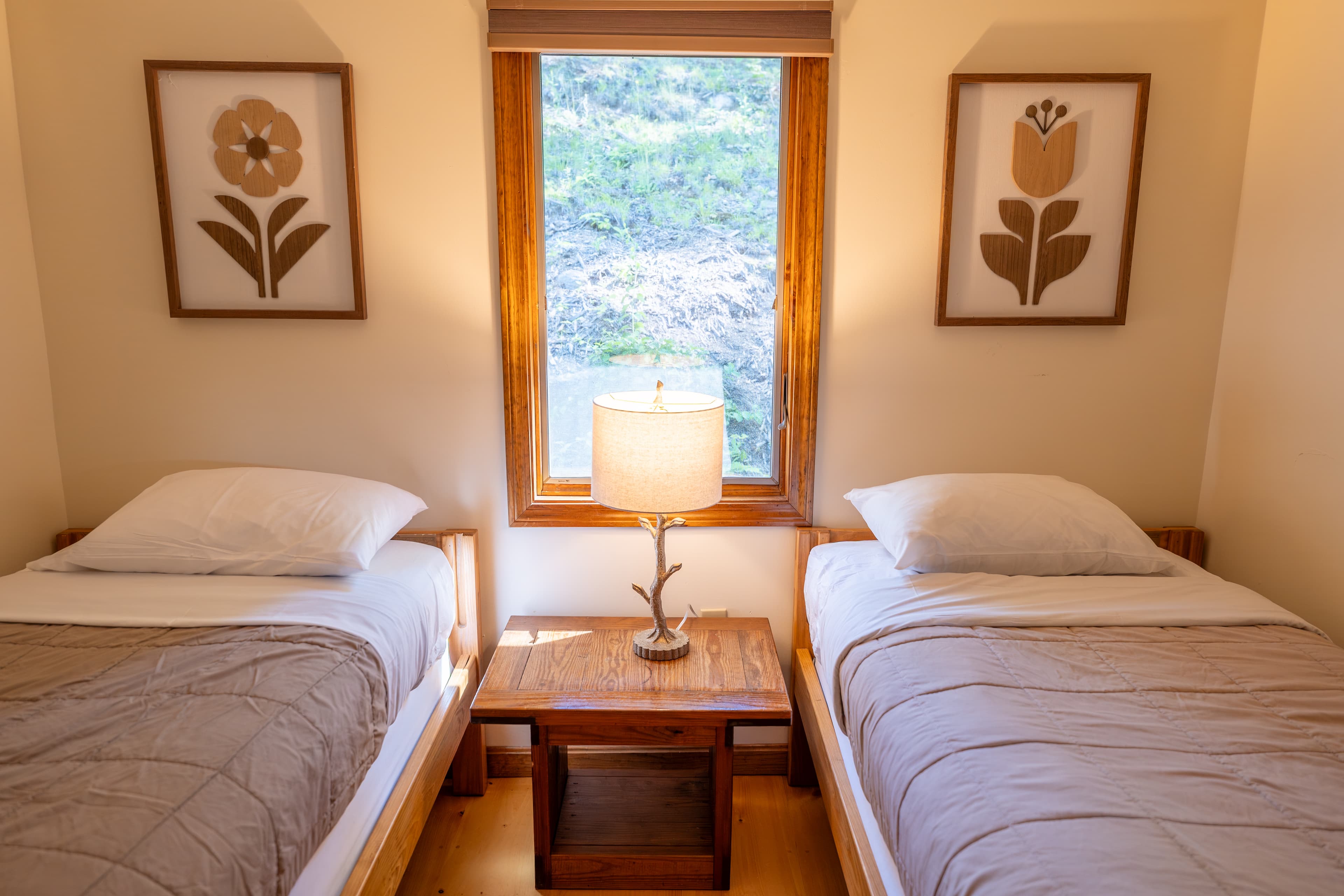 A simple cabin bedroom featuring two twin beds with patterned covers, separated by a wooden nightstand with a lamp under a curtained window.