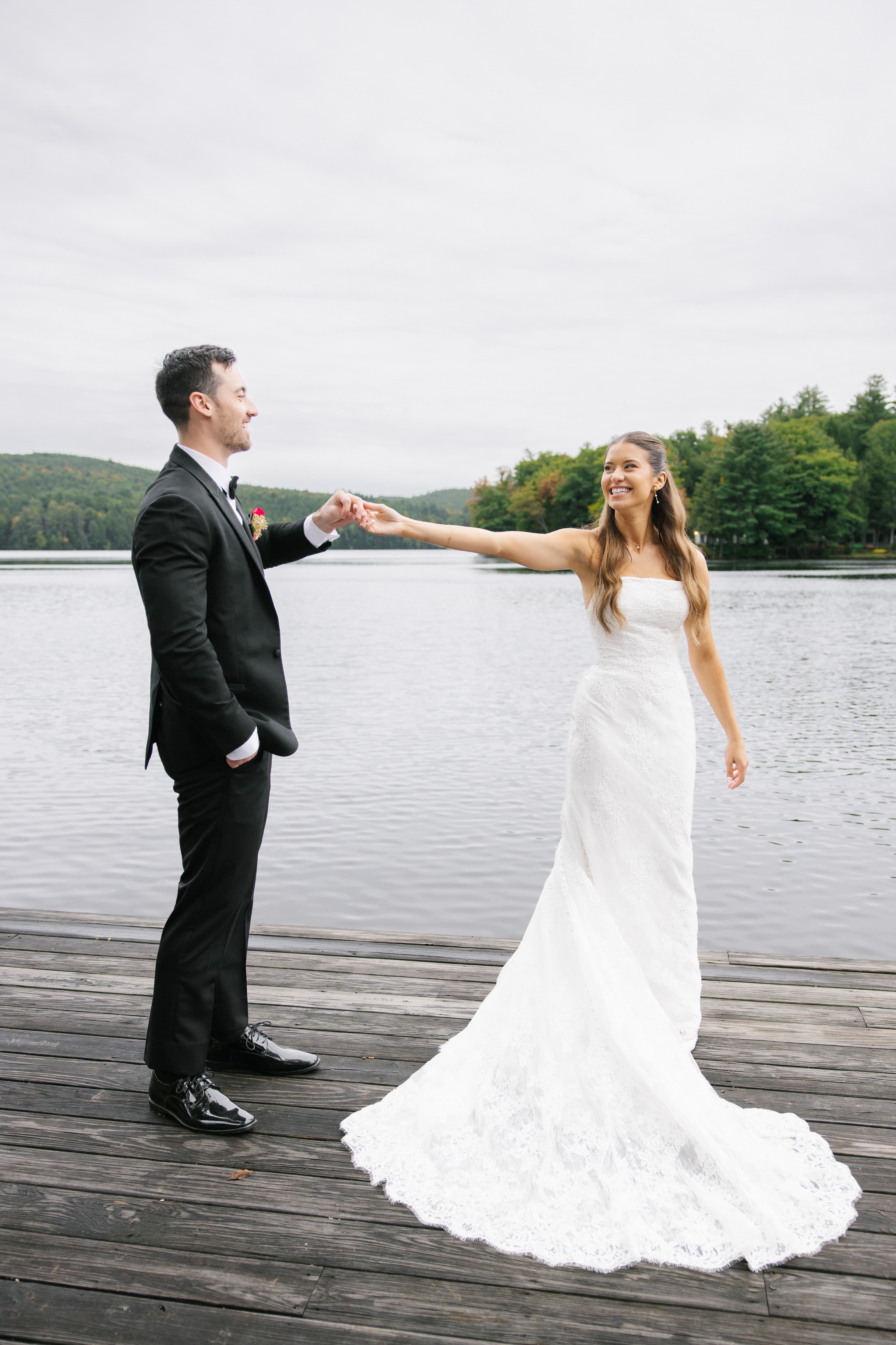 A bride in a lace gown holds hands with a man in a suit by a serene lake.