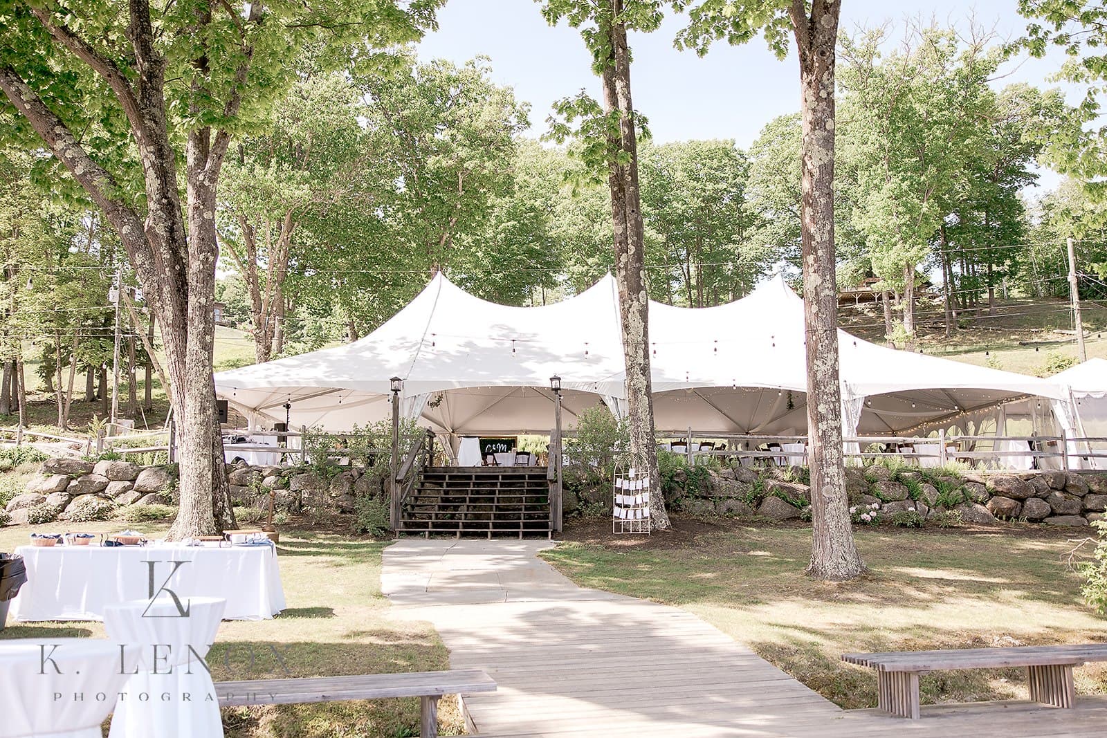 A large white tent is set up among trees, with a wooden walkway leading to it.