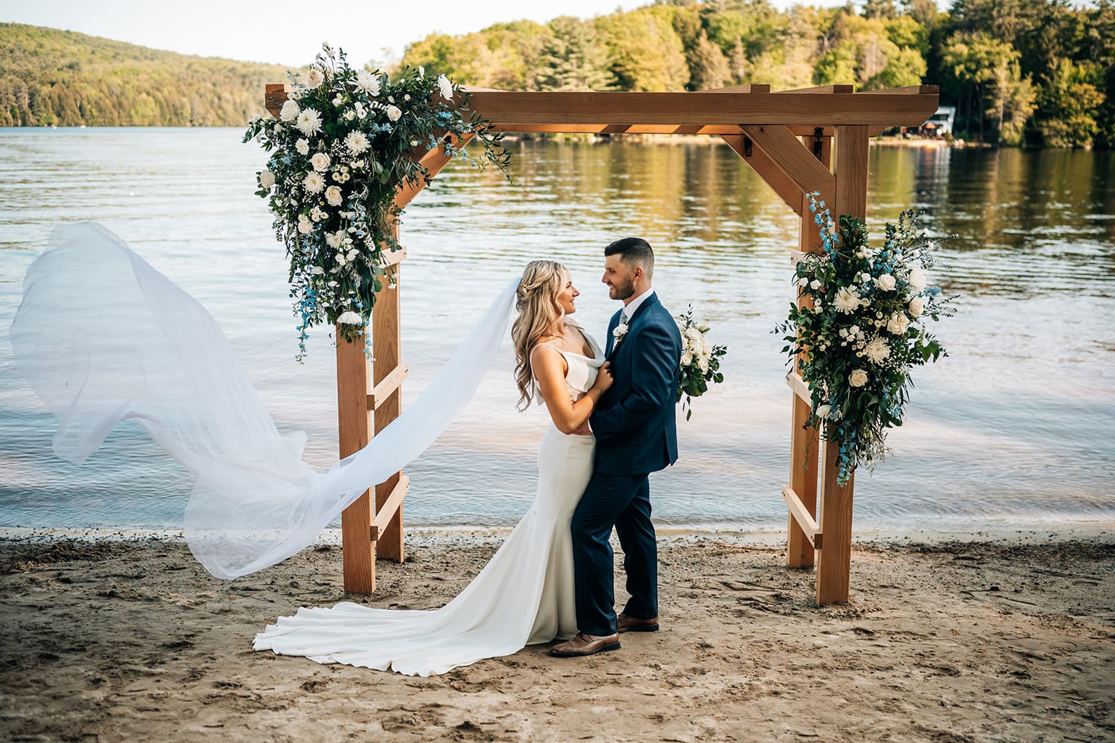 A bride and groom share a romantic moment under a floral arch by the water.
