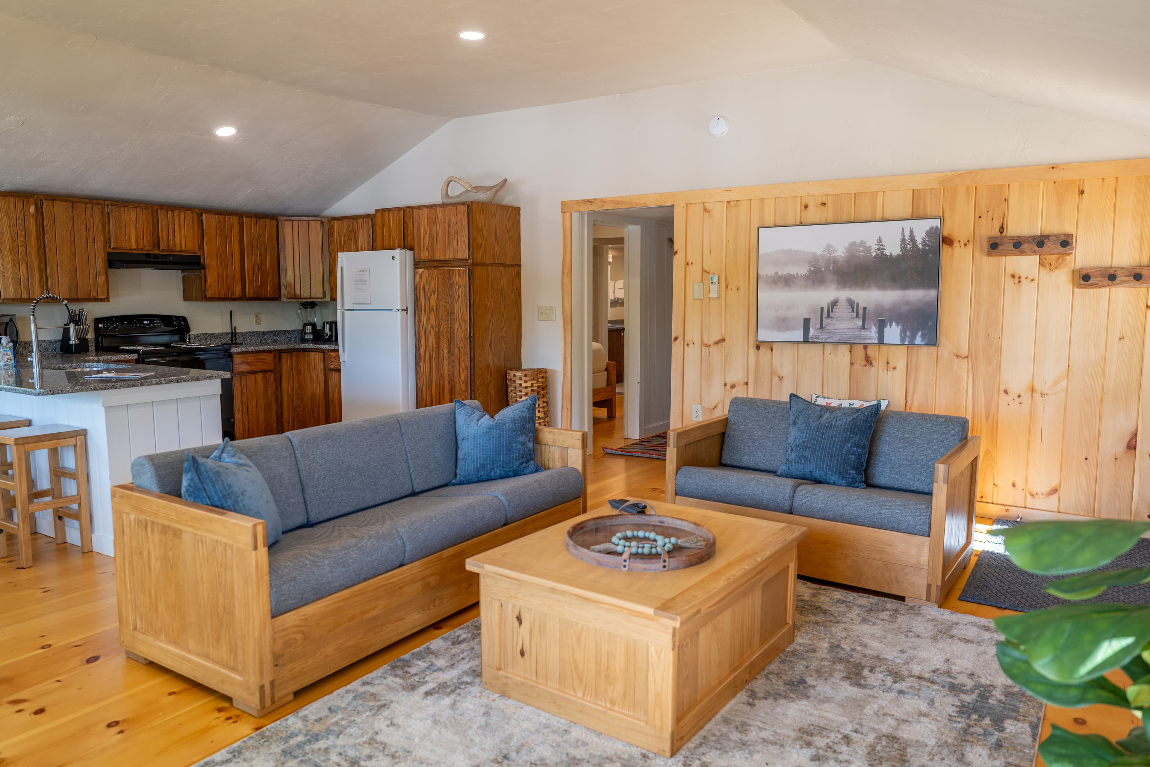 An open-plan cabin great room with a vaulted ceiling, featuring a living area with a plaid sofa and an open-concept kitchen with wood cabinets.