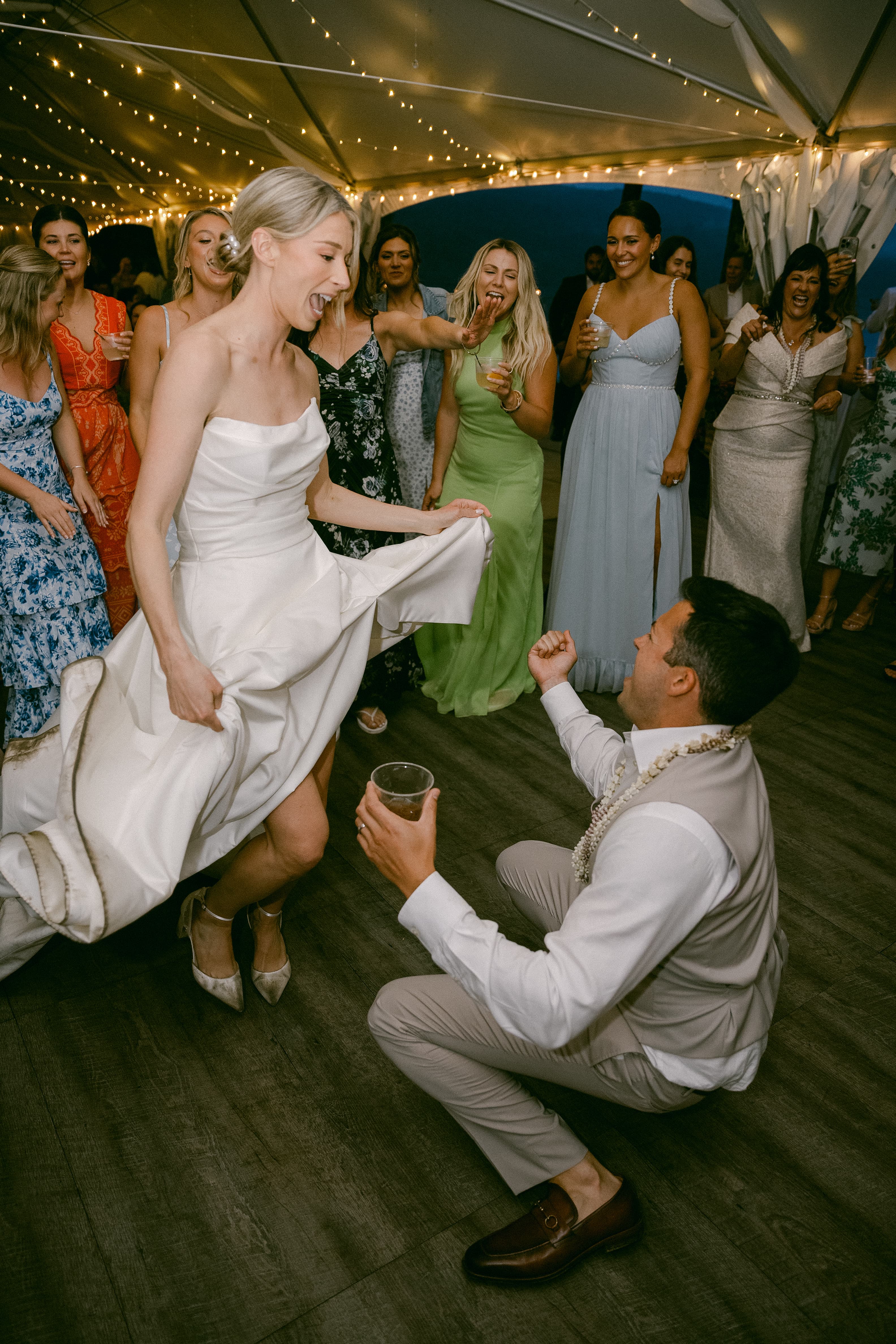 A bride dances joyfully while a man kneels, surrounded by guests celebrating at a festive event.